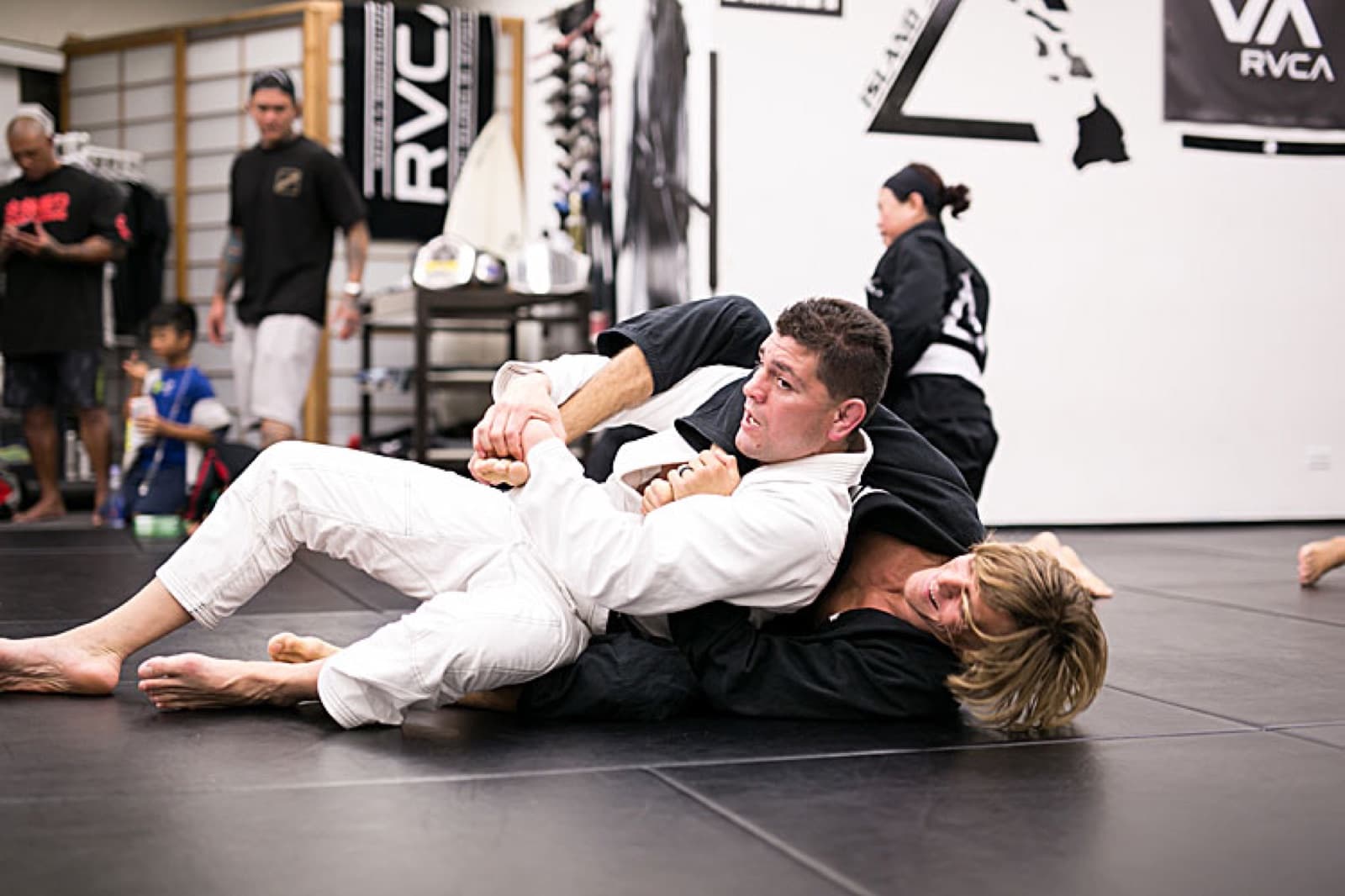 Two adults grappling on the black mats during an adult class — one in a white gi, one in a black gi