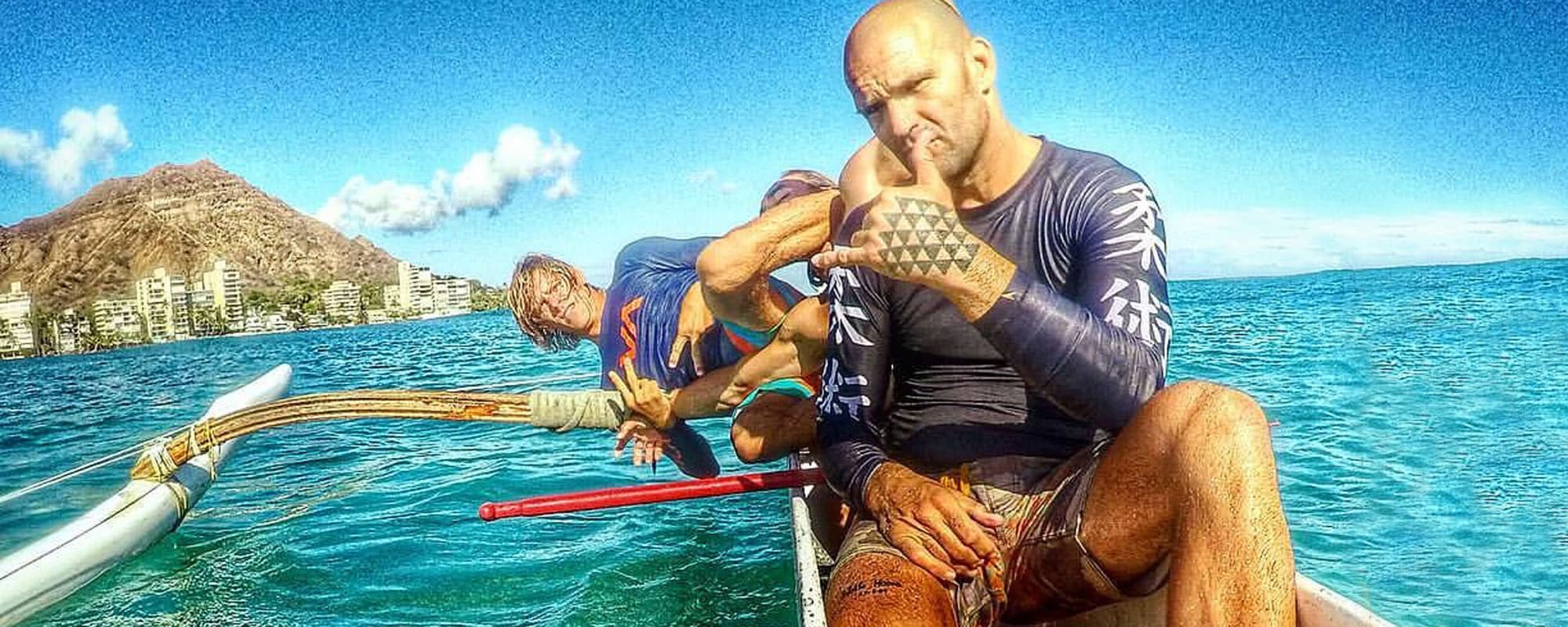 Two Island Jiu-Jitsu members throwing shakas from an outrigger canoe off Waikīkī, with Diamond Head behind them