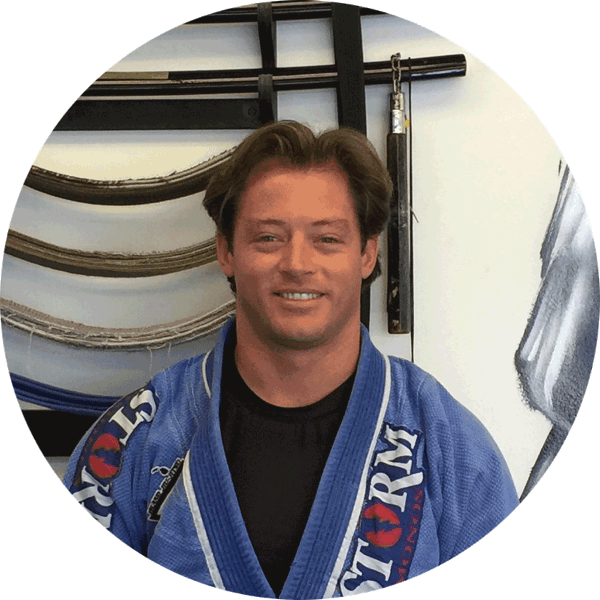 Dan Chipchase smiling in a blue gi in front of a rack of belts