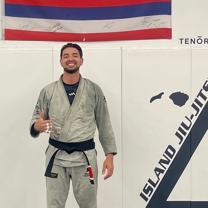 Professor Elijah Edwards in a grey gi and black belt in front of the Hawaiʻi flag and the Island Jiu-Jitsu logo wall