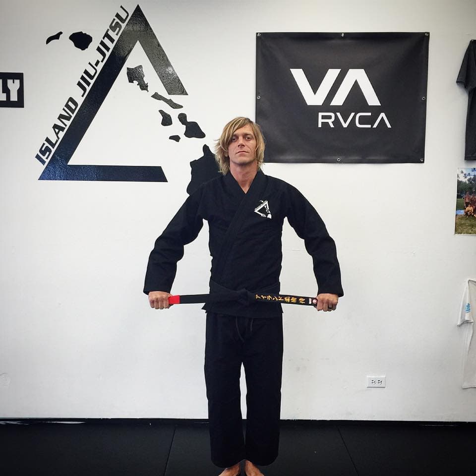 A coach wearing the black Island Jiu-Jitsu team gi in front of the IJJ triangle logo on the dojo wall