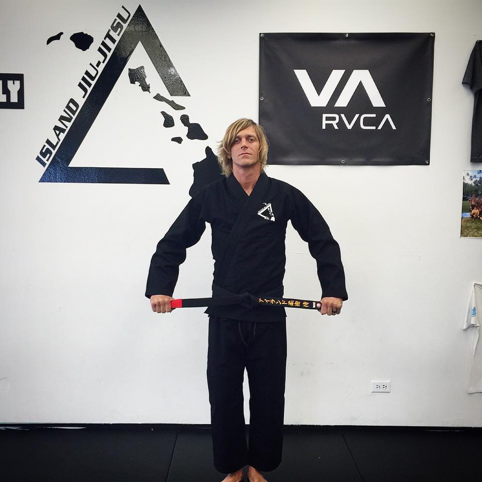 A coach wearing the black Island Jiu-Jitsu team gi, holding a black belt across his waist in front of the IJJ triangle logo painted on the dojo wall