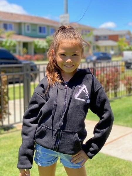 A keiki student wearing the black IJJ zip hoodie with the small white logo on the chest, standing on a sunny sidewalk