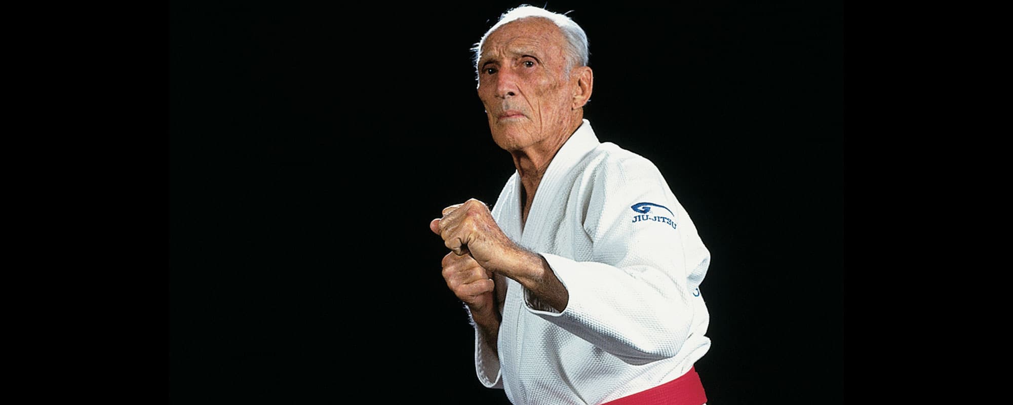 Helio Gracie in a white gi and red belt, fists raised in a fighting stance against a black background