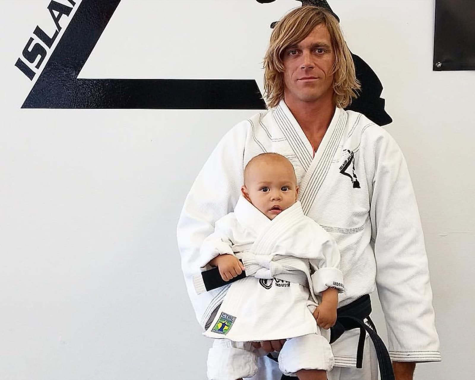 Jason Vigil in a white gi and black belt, holding a baby in a tiny white gi, in front of the Island Jiu-Jitsu logo wall